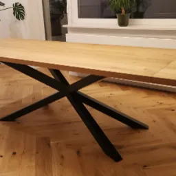 Rozkładany stół z blatem z jasnego drewna dębowego i czarnymi, metalowymi nogami w kształcie gwiazdy, na podłodze ułożonej w jodełkę.