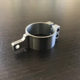 Metalowy uchwyt z otworem na śrubę i dwoma otworami montażowymi, obrobiony na gładko, leżący na ciemnym, drewnianym blacie.