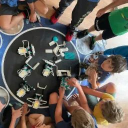 Grupa dzieci siedzi wokół okrągłego stołu z motywem kosmosu, budując roboty z klocków. Jeden z robotów ma wyświetlacz z włączonymi diodami. Dzieci w różnym wieku i ubraniach skupione na zadaniu.