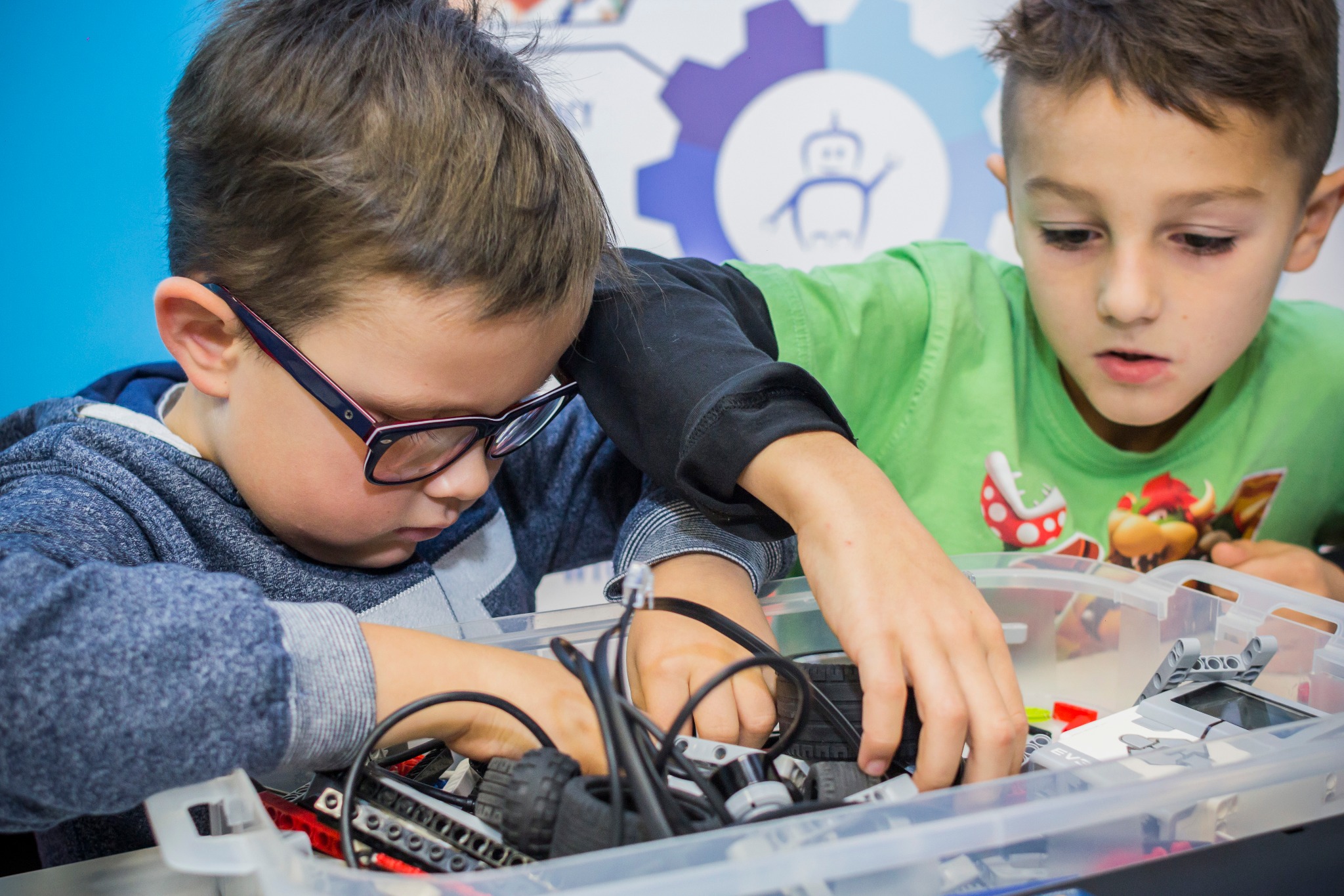 Dwóch chłopców skupionych na budowaniu z klocków w przezroczystym pojemniku; w tle logo przedstawiające robota na tle zębatki.