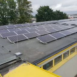 Rząd paneli słonecznych na płaskim dachu budynku z ż&oacute;łtymi elementami architektonicznymi, widok z g&oacute;ry na tle drzew i pochmurnego nieba.