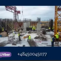 Widok z góry na plac budowy z pracownikami w kamizelkach odblaskowych, dźwigami i zbrojeniami, w tle widoczne morze.