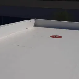 Biała, płaska powierzchnia dachu z widocznym czerwonym uszczelnieniem wokół elementu mocującego, białe obrzeże dachu w tle.