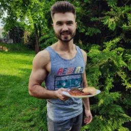 Uśmiechnięty mężczyzna w szarej koszulce na ramiączkach z nadrukiem, prezentuje talerz z posiłkiem wysokobiałkowym na tle zielonego ogrodu i krzewu.