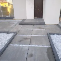 Nowoczesne podejście do domu wyłożone dużymi, szarymi płytami betonowymi oddzielonymi pasami białego grysu, z obrzeżami z kostki brukowej i rabatami wypełnionymi białym kamieniem.