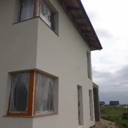 Dwupiętrowy dom w trakcie prac elewacyjnych, z oknami zabezpieczonymi folią, świeżo otynkowaną elewacją w jasnym odcieniu i widocznymi elementami dachu z brązową dachówką. W tle widoczne inne domy...