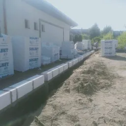 Wznoszenie ścian fundamentowych z bloczków Solbet, widoczne palety z materiałem budowlanym na placu budowy.