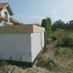 Wznoszenie ścian z białych bloczków betonu komórkowego Ytong, z widocznym deskowaniem z płyty OSB na górze, oraz izolacją przeciwwilgociową fundamentu, na tle ogrodu i fragmentu domu jednorodzinnego.