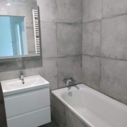 PRZEROBOTA Kamil Przesław - Wykończona łazienka z szarymi płytkami, białą umywalką z szafką i wanną. Widoczne lustro z grzejnikiem. Minimalistyczny design, nowoczesne wykończenie.