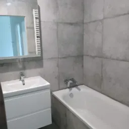 Wykończona łazienka z szarymi płytkami, białą umywalką z szafką i wanną. Widoczne lustro z grzejnikiem. Minimalistyczny design, nowoczesne wykończenie.