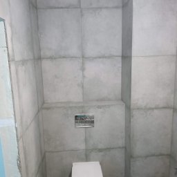 PRZEROBOTA Kamil Przesław - Nowoczesna toaleta z szarymi, kwadratowymi płytkami imitującymi beton, podwieszana miska WC i chromowany przycisk spłukujący. Widoczny fragment niedokończonej ściany.