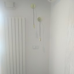 PRZEROBOTA Kamil Przesław - Biała ściana po odświeżeniu w Nowej Woli, widoczne dwa złote kinkiety z okrągłymi mocowaniami, biały grzejnik panelowy oraz klimatyzator pod sufitem.