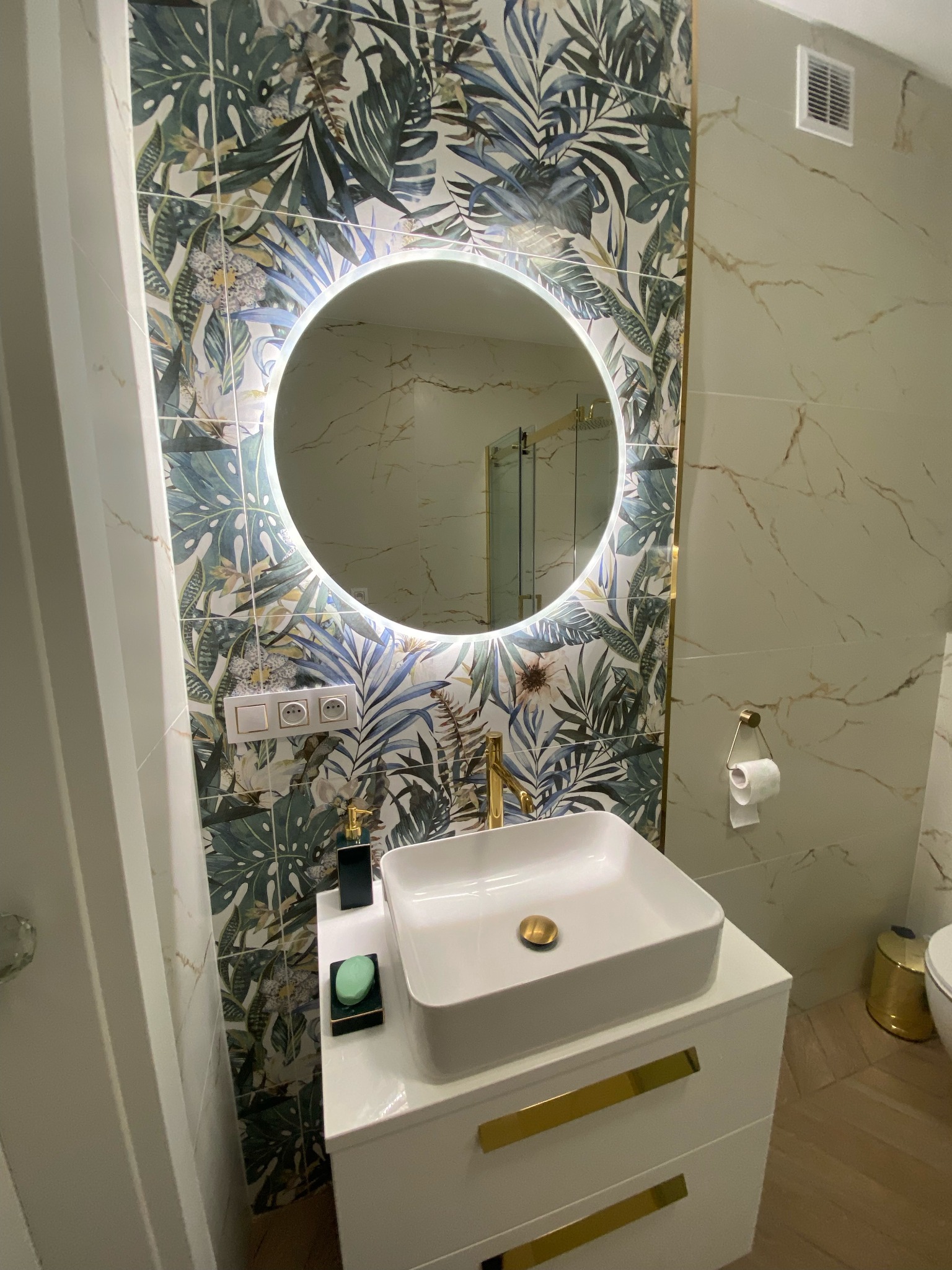 Nowoczesna łazienka z umywalką nablatową, okrągłym lustrem z podświetleniem LED, baterią w kolorze złota i płytkami z motywem roślinnym.