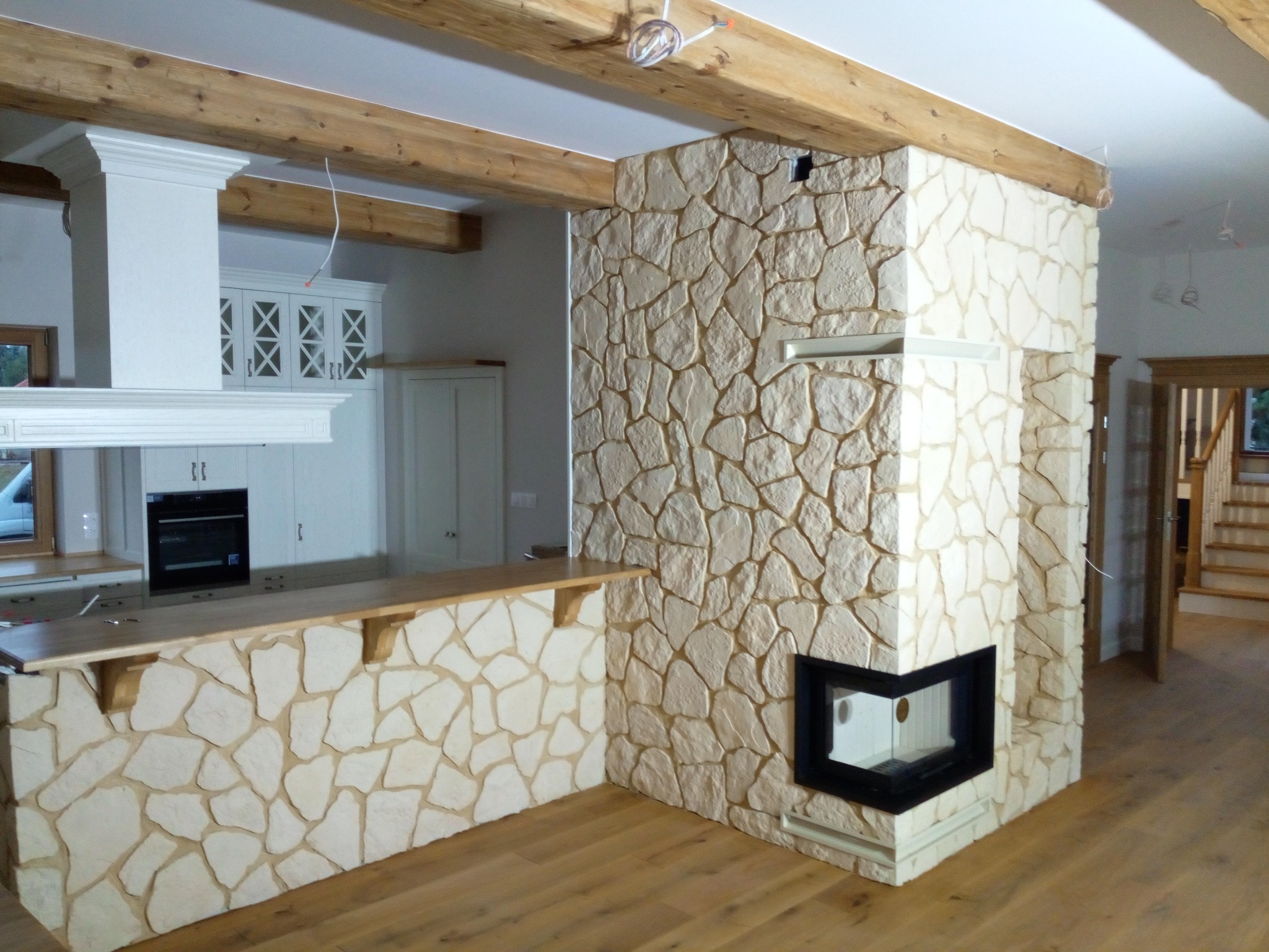 Wykończona ściana z kamienia dekoracyjnego w salonie z kominkiem narożnym i drewnianą podłogą, widoczne drewniane belki stropowe i fragment zabudowy kuchennej w tle.