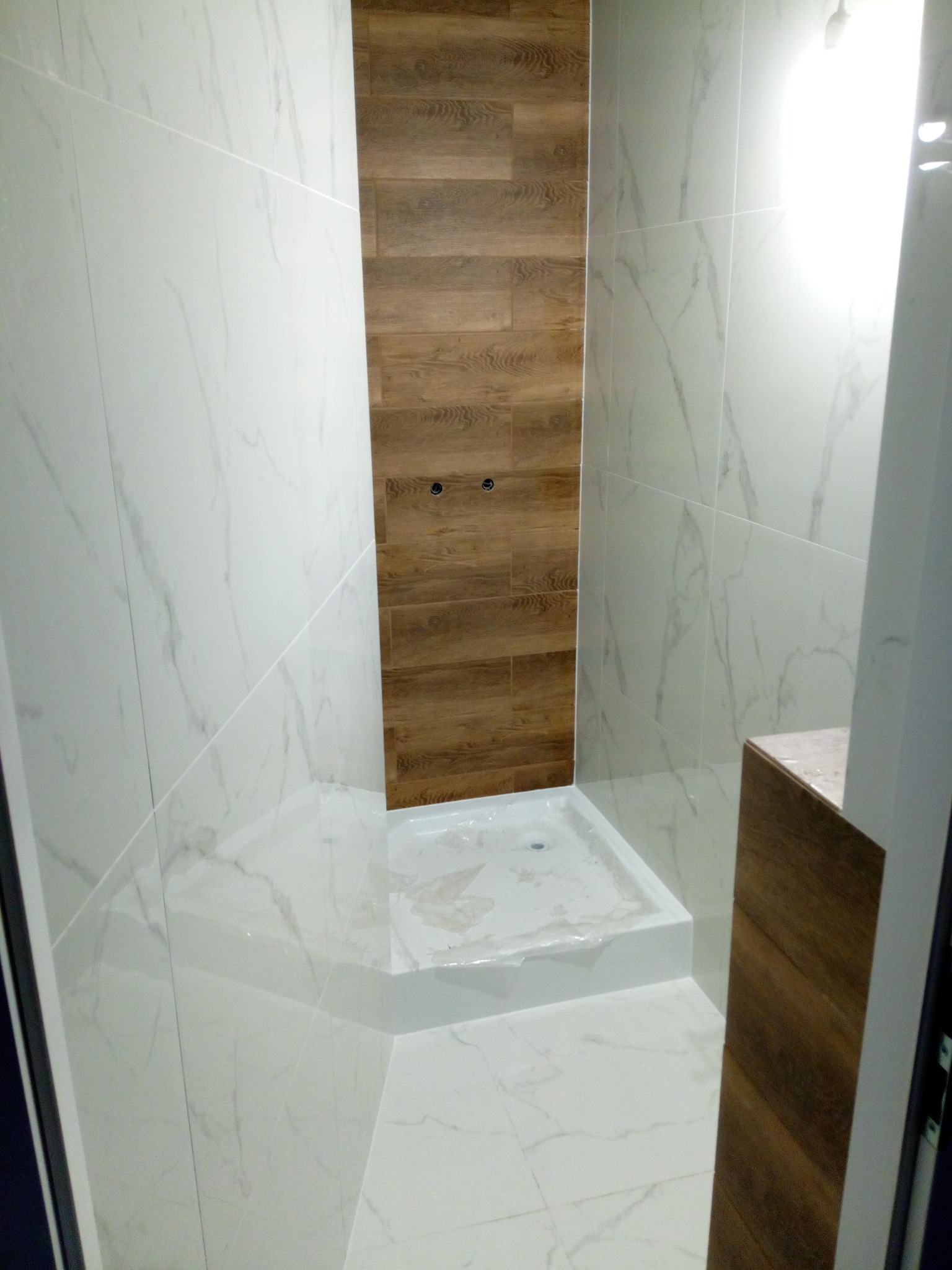 Wykończona kabina prysznicowa z białymi płytkami imitującymi marmur i panelem ściennym imitującym drewno, widoczne elementy instalacji.