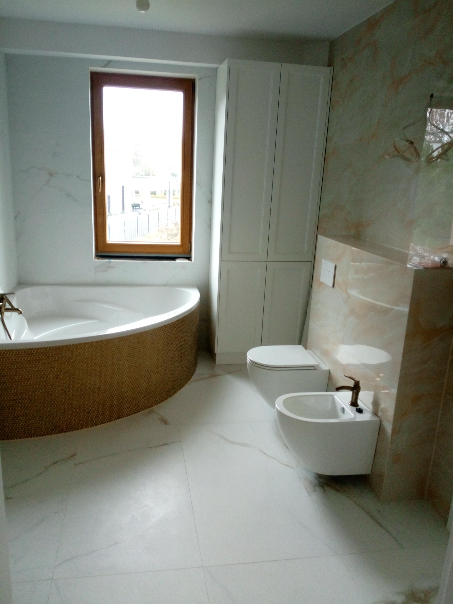 Wykończona łazienka z wanną z mozaiką, białą armaturą i płytkami imitującymi marmur. Okno z drewnianą ramą.