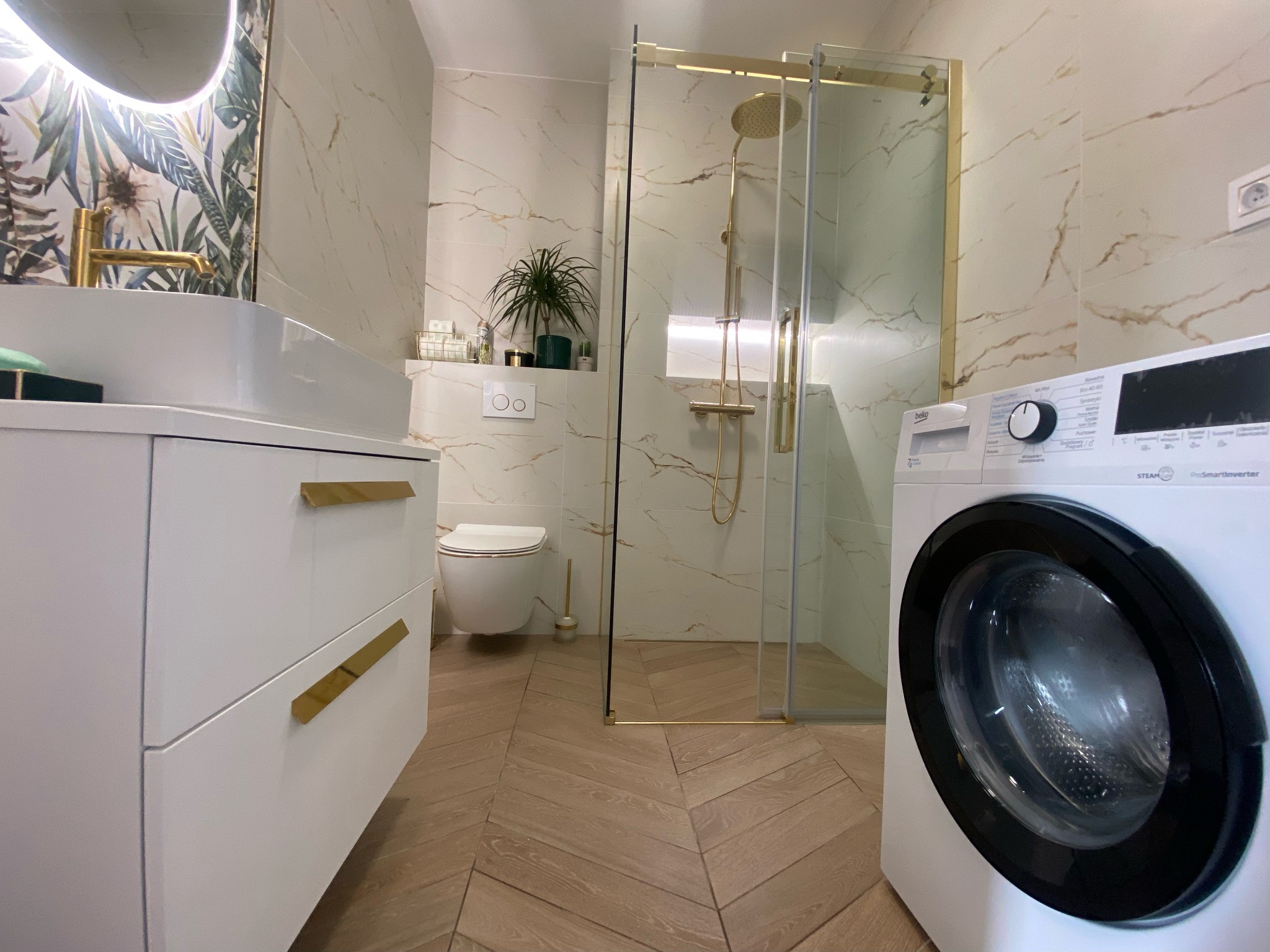 Nowoczesna łazienka z kabiną prysznicową w złotej ramie, umywalką na szafce z białymi frontami i złotymi uchwytami, pralką oraz toaletą w tle, wykończona płytkami imitującymi marmur i parkietem...