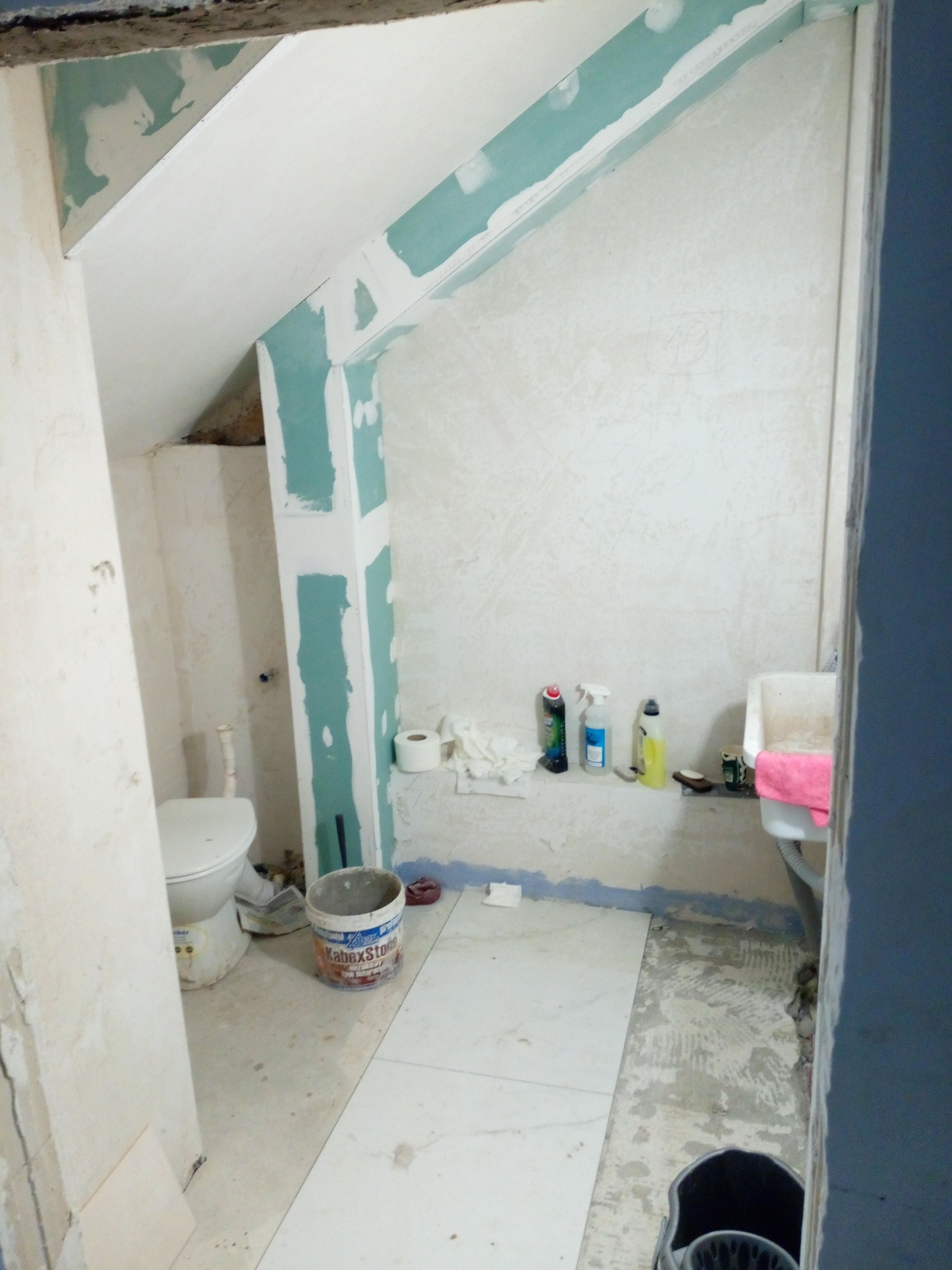 Wnętrze łazienki w trakcie remontu: surowe ściany z płyt gipsowo-kartonowych, częściowo położone płytki podłogowe, widoczna toaleta i umywalka, oraz akcesoria malarskie i środki czystości na półce.