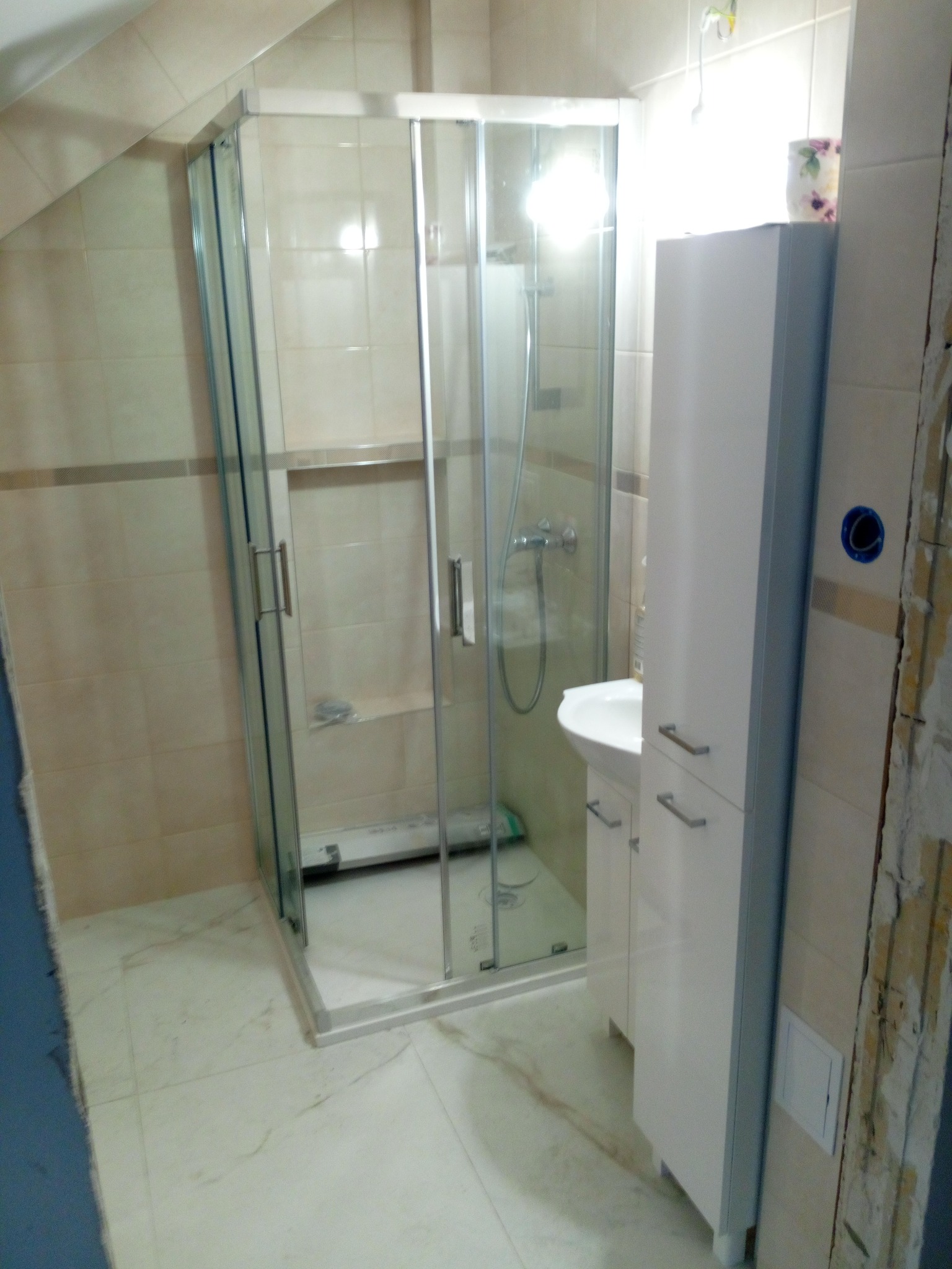 Wyremontowana łazienka z narożną kabiną prysznicową z przezroczystego szkła, białą szafką z umywalką i fragmentem ściany w trakcie wykończenia z widoczną instalacją elektryczną.