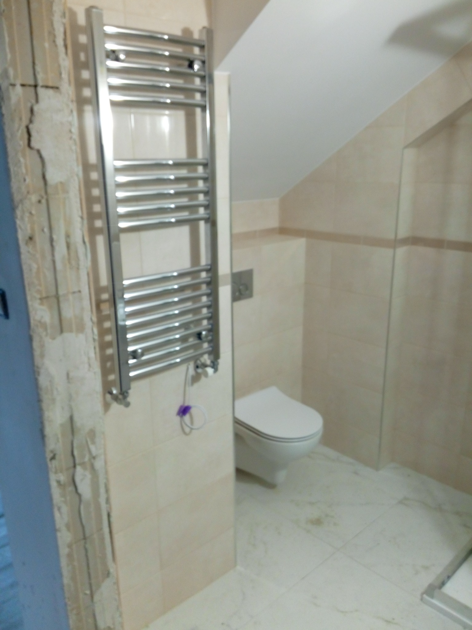 Wnętrze łazienki w trakcie remontu, widoczny grzejnik drabinkowy na ścianie z beżowych płytek, obok ściana z odsłoniętym fragmentem muru i instalacją elektryczną, w tle podwieszana toaleta i kabina...