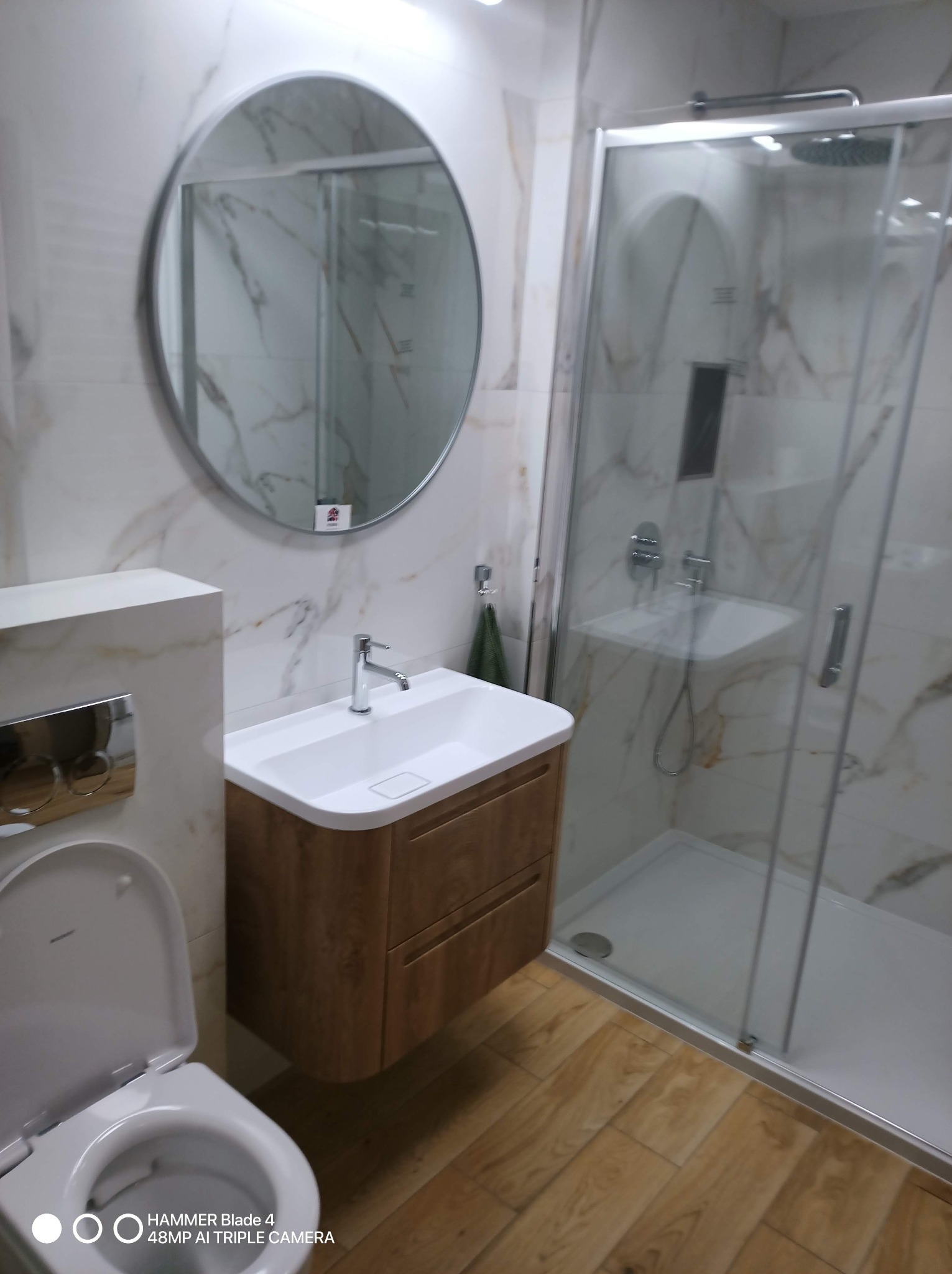 Wykończona łazienka z kabiną prysznicową, umywalką z drewnianą szafką i okrągłym lustrem. Marmurowe płytki na ścianach. Jasna podłoga imitująca drewno.