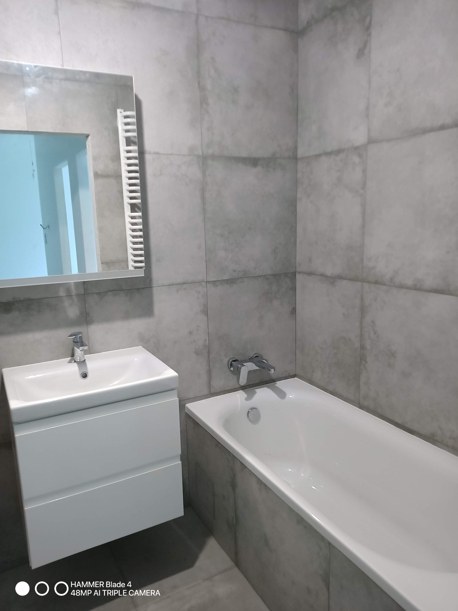 Wykończona łazienka z szarymi płytkami, białą umywalką z szafką i wanną. Widoczne lustro z grzejnikiem. Minimalistyczny design, nowoczesne wykończenie.