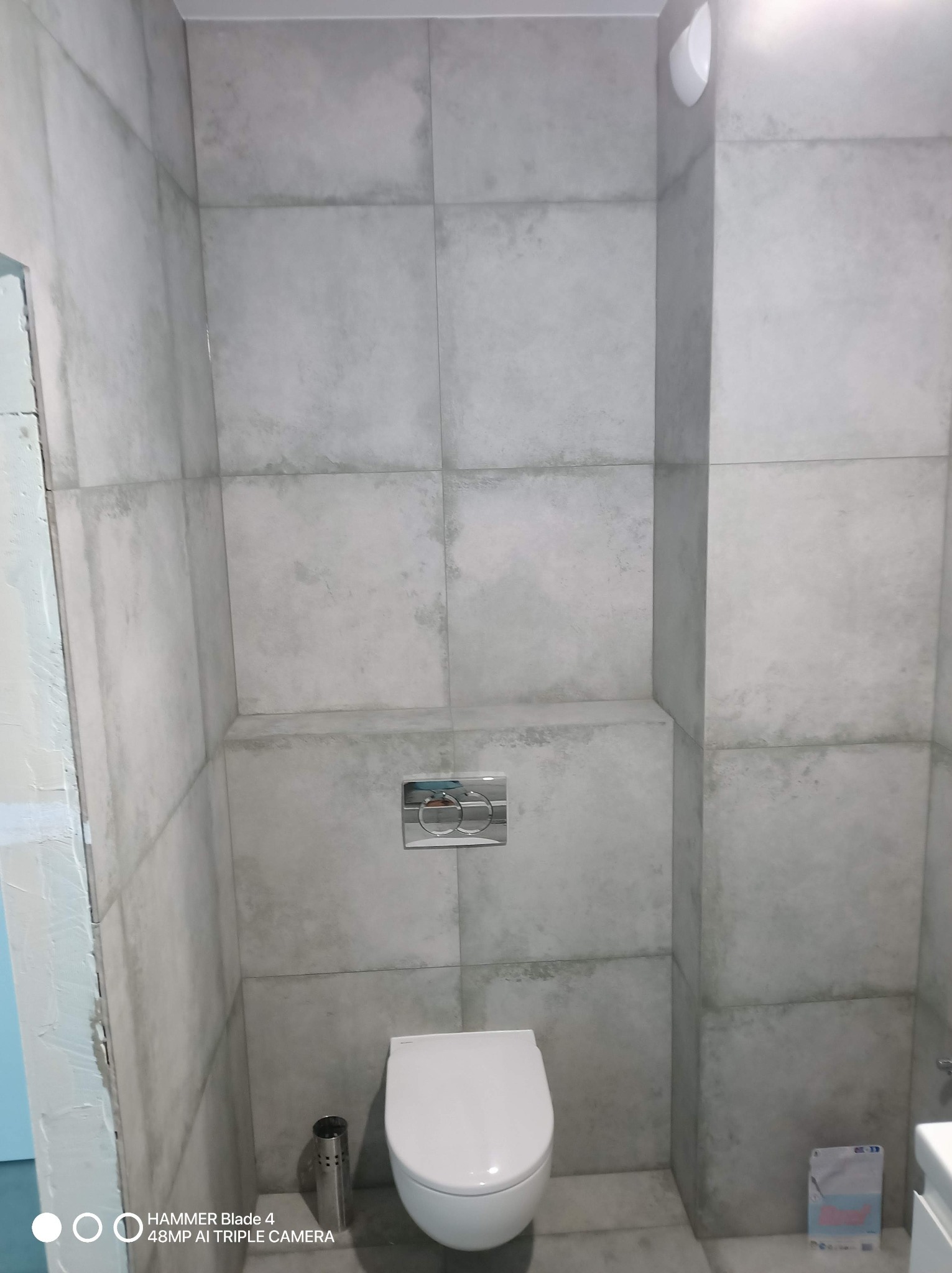 Nowoczesna toaleta z szarymi, kwadratowymi płytkami imitującymi beton, podwieszana miska WC i chromowany przycisk spłukujący. Widoczny fragment niedokończonej ściany.