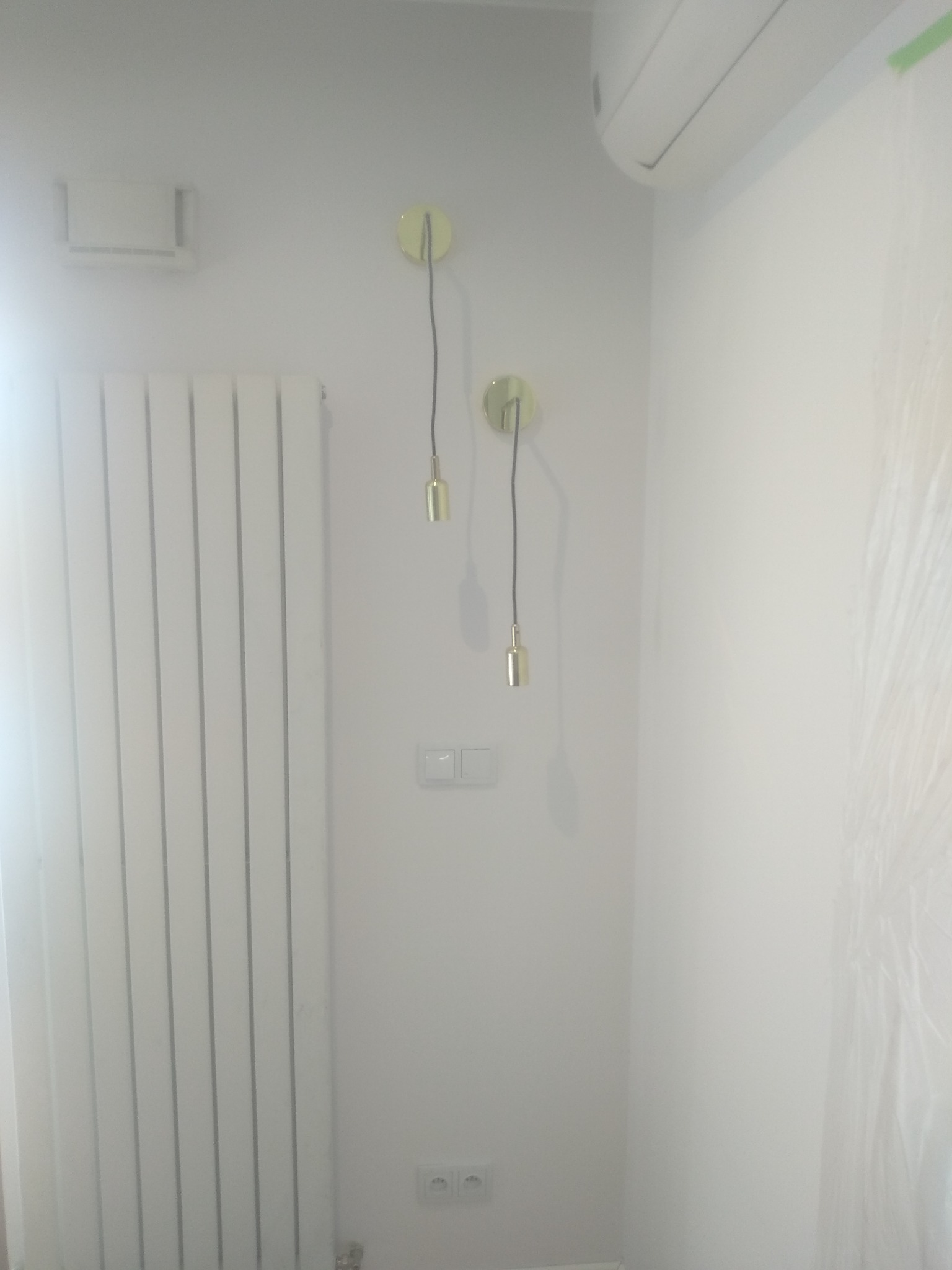 Biała ściana po odświeżeniu w Nowej Woli, widoczne dwa złote kinkiety z okrągłymi mocowaniami, biały grzejnik panelowy oraz klimatyzator pod sufitem.