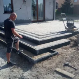 Pracownik w trakcie układania schodów i tarasu z kostki, widoczna zagęszczarka w tle, częściowo ułożony taras z obramowaniem, etap prac ziemnych.