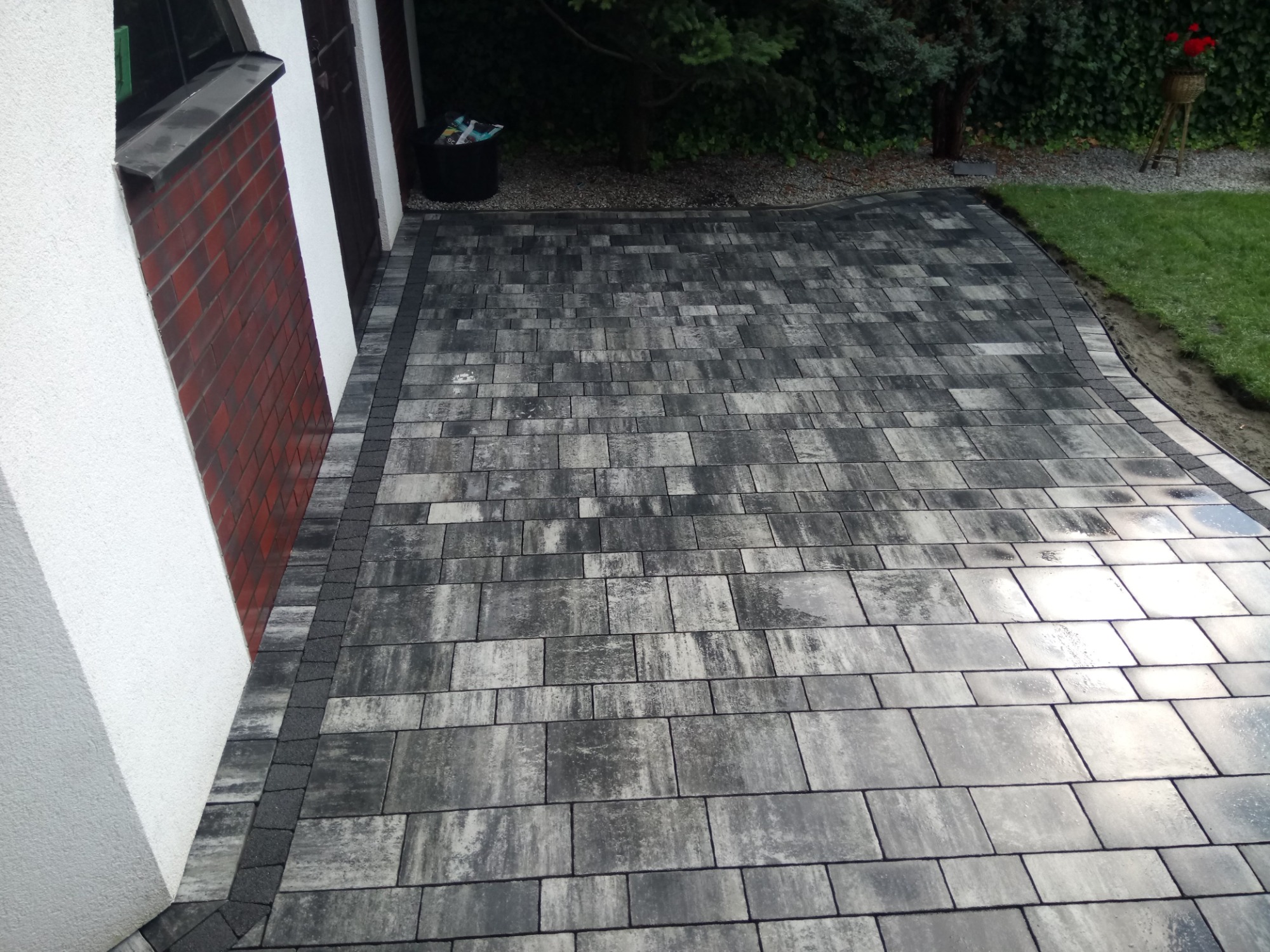 Ułożona szara kostka brukowa tworząca ścieżkę obok domu z ceglaną ścianą i trawnikiem w tle.