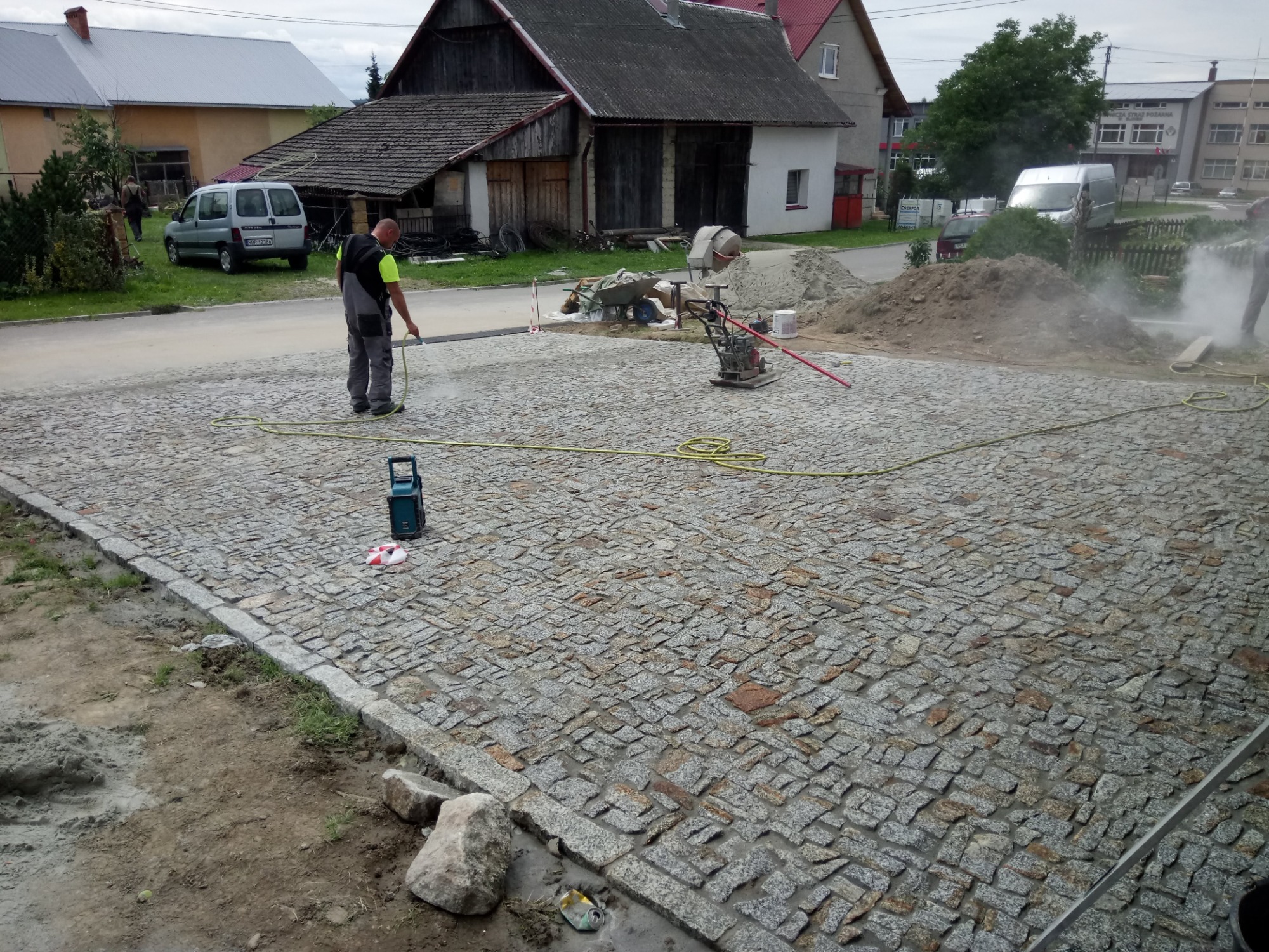 Układanie kostki granitowej: robotnik spryskuje wodą nowo ułożoną nawierzchnię z kostki granitowej o nieregularnym kształcie, widoczna ubijarka, betoniarka, hałda piasku i okoliczna zabudowa.