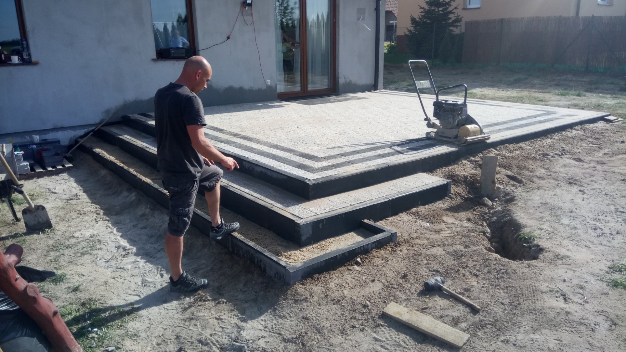 Pracownik w trakcie układania schodów i tarasu z kostki, widoczna zagęszczarka w tle, częściowo ułożony taras z obramowaniem, etap prac ziemnych.