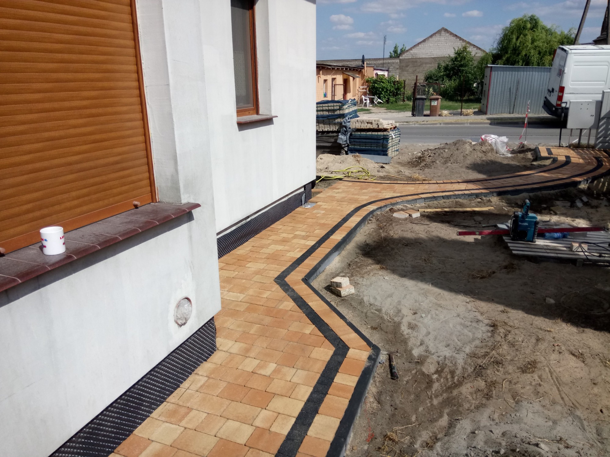 Zakręcony chodnik z żółtej kostki brukowej z czarnym obramowaniem, częściowo ułożony, z widocznymi narzędziami i stertą kostki w tle, obok białego budynku z brązowymi roletami.