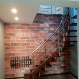 Schody drewniane z metalową balustradą na tle ściany z cegieł, oświetlone punktowo, z dekoracyjnym elementem imitującym kratę.