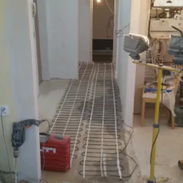 Instalacja ogrzewania podłogowego w korytarzu podczas prac wykończeniowych, widoczne narzędzia, materiały budowlane i lampa halogenowa na statywie.