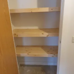 Wnętrze szafy wnękowej z trzema surowymi, sosnowymi półkami, widoczna surowa podłoga w trakcie wykończenia.