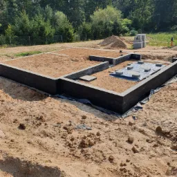Fundament z bloczków betonowych, częściowo wypełniony piaskiem, na działce z widocznymi drzewami w tle.