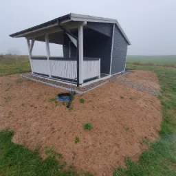 Niewielka, ciemnoszara konstrukcja z białymi balustradami, prawdopodobnie altana lub domek narzędziowy, umieszczona na nasypie z piasku i kamieni na tle zamglonego krajobrazu polnego.