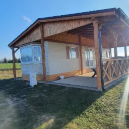 Gotowy, parterowy dom z elewacją z paneli, drewnianym zadaszeniem nad tarasem i ozdobną, krzyżową balustradą, na tle zielonego trawnika i błękitnego nieba.