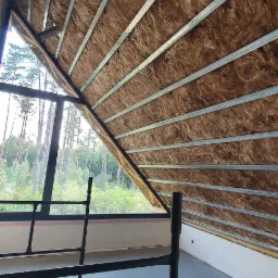 Wnętrze poddasza z widoczną izolacją z wełny mineralnej między metalowymi profilami, rusztowanie w pomieszczeniu, okno z widokiem na las.