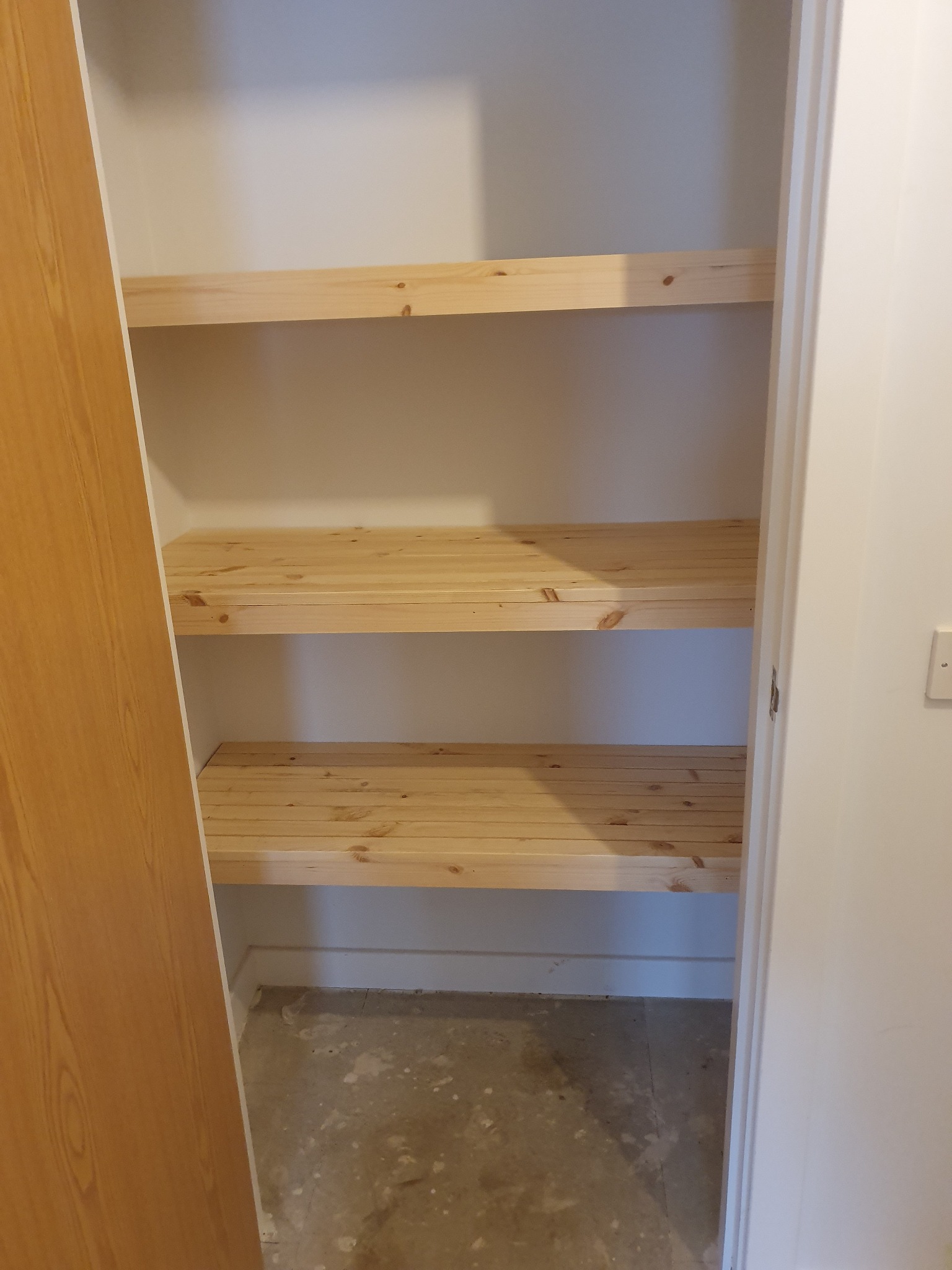 Wnętrze szafy wnękowej z trzema surowymi, sosnowymi półkami, widoczna surowa podłoga w trakcie wykończenia.