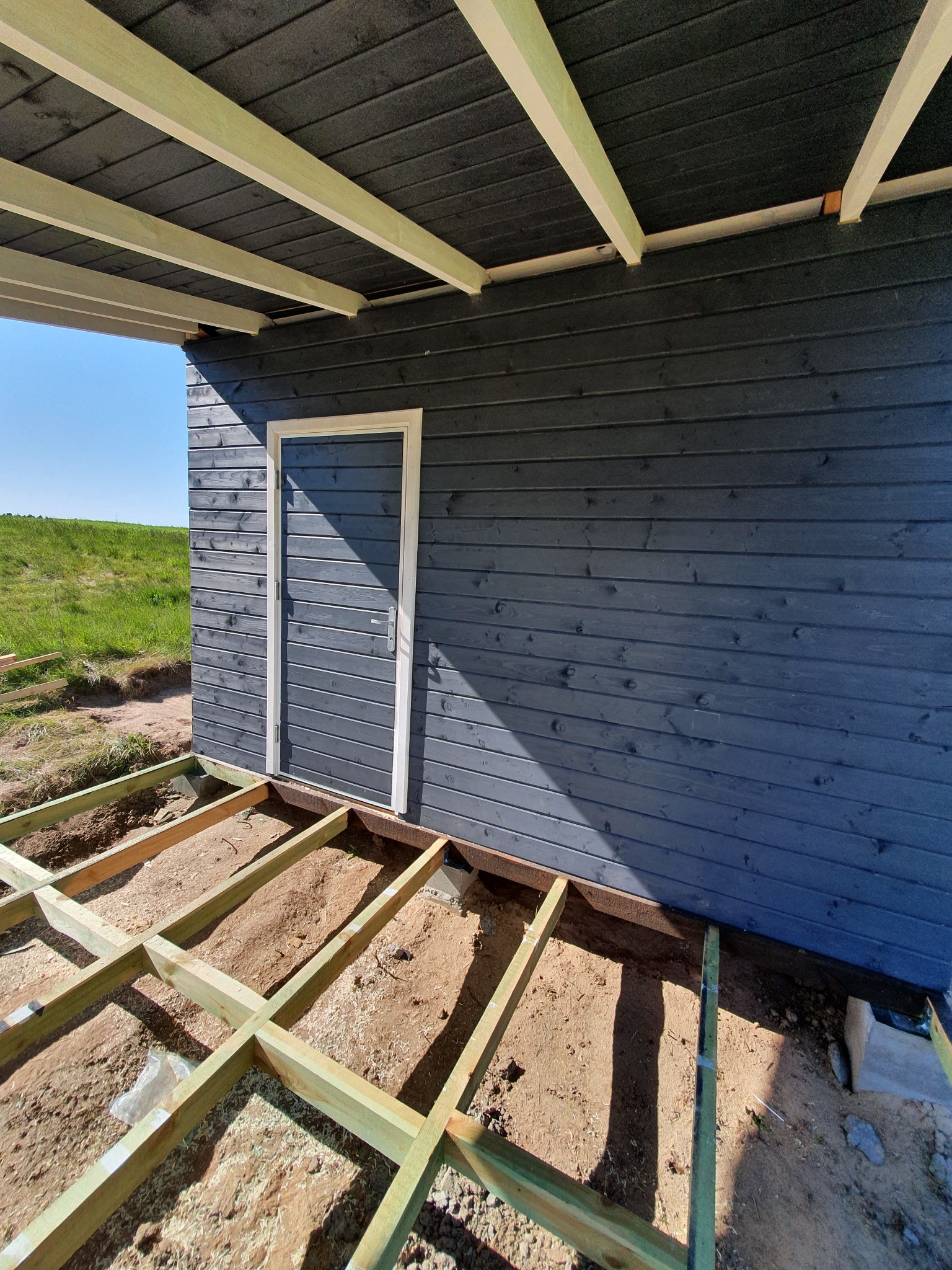 Budowa domu z drewnianą elewacją: widok na fundament tarasu, ściany z ciemnego drewna i białe obramowanie drzwi.