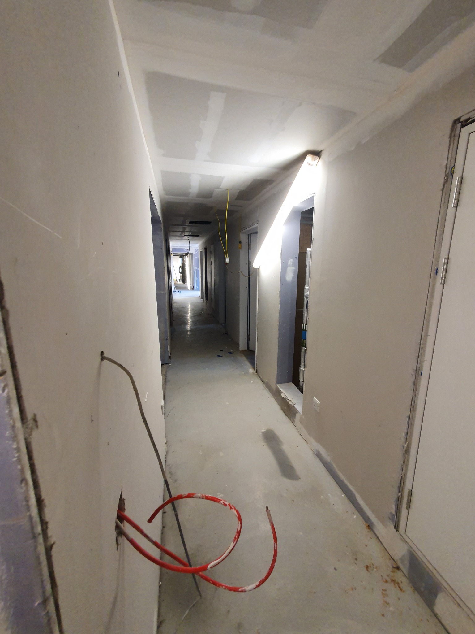 Korytarz w trakcie prac wykończeniowych: ściany przygotowane pod malowanie, widoczne przewody elektryczne i rury grzewcze wystające ze ściany, sufit z płyt kartonowo-gipsowych w trakcie szpachlowania