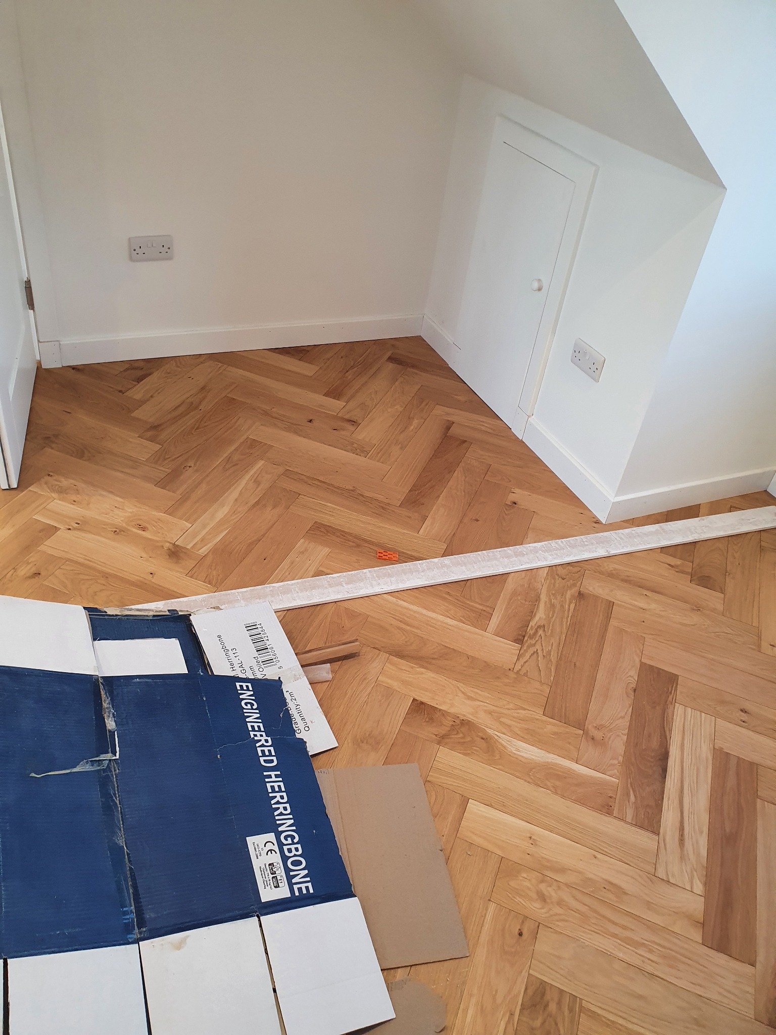 Ukończona podłoga z parkietu ułożonego w jodełkę, widoczny fragment kartonu po panelach i listwa przypodłogowa przed montażem w narożniku pokoju.