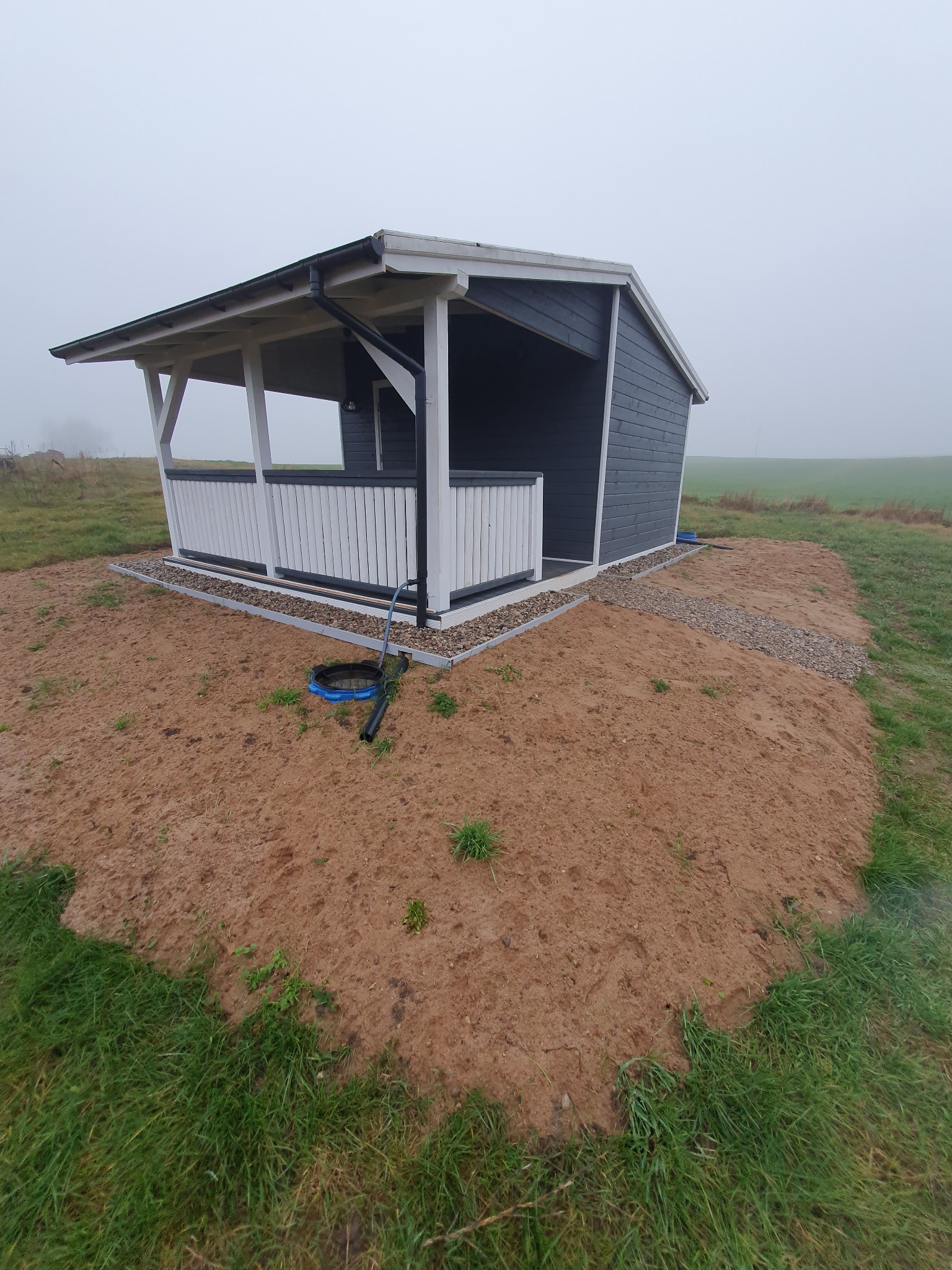 Niewielka, ciemnoszara konstrukcja z białymi balustradami, prawdopodobnie altana lub domek narzędziowy, umieszczona na nasypie z piasku i kamieni na tle zamglonego krajobrazu polnego.