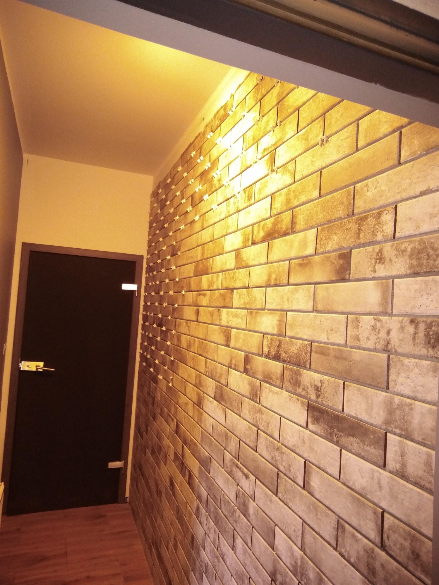 Ściana wyłożona cegiełkami w trakcie remontu, widoczne krzyżyki dystansowe, w tle ciemne drzwi i fragment podłogi.