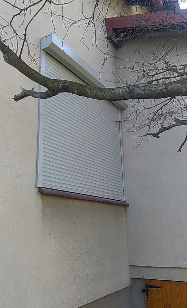 Zamknięta, zewnętrzna roleta okienna w kolorze jasnoszarym, widoczna na tle beżowej ściany budynku i gałęzi drzewa.