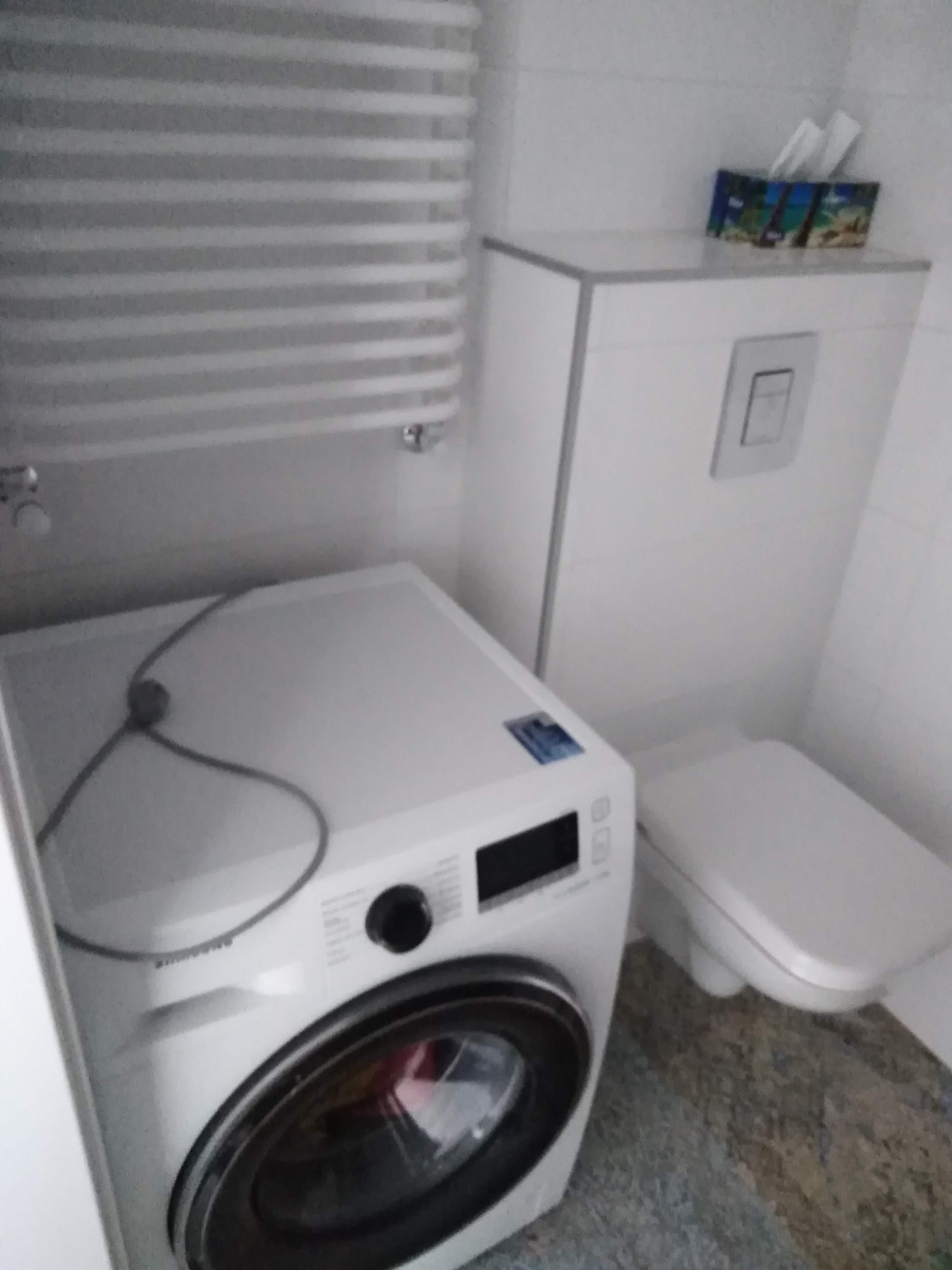 Biała pralka Samsung w łazience, obok toaleta typu Geberit z zabudową, na ścianie grzejnik drabinkowy, a na zabudowie toalety pudełko chusteczek.