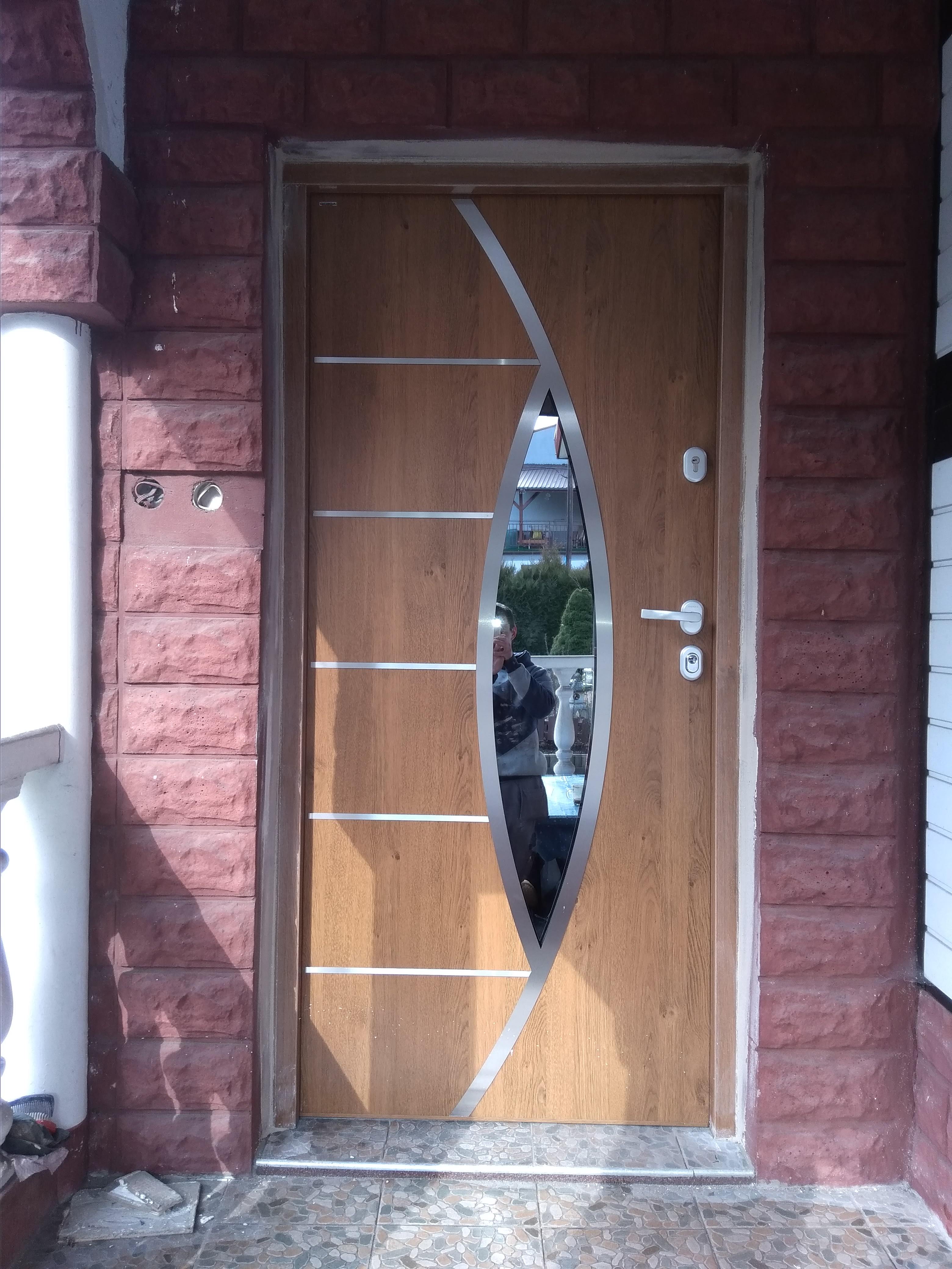 Drewniane drzwi wejściowe z dekoracyjnymi, srebrnymi wstawkami i owalnym przeszkleniem, zamontowane w ceglanej wnęce. W szybie odbija się osoba robiąca zdjęcie.