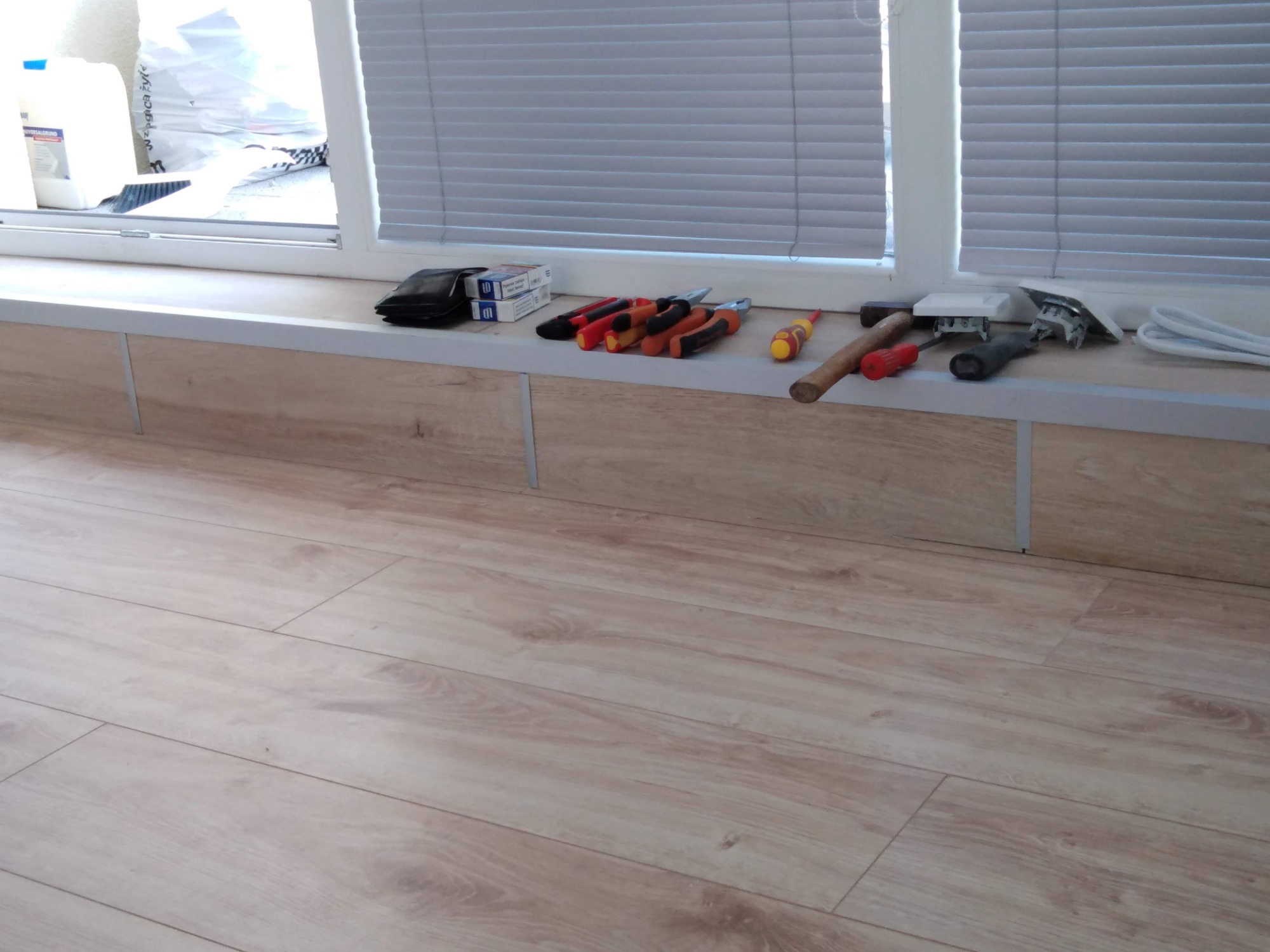 Narzędzia, w tym śrubokręty, kombinerki, młotek i przewody elektryczne, ułożone na parapecie okiennym z drewnianą okładziną, widok z bliska na podłogę z paneli.